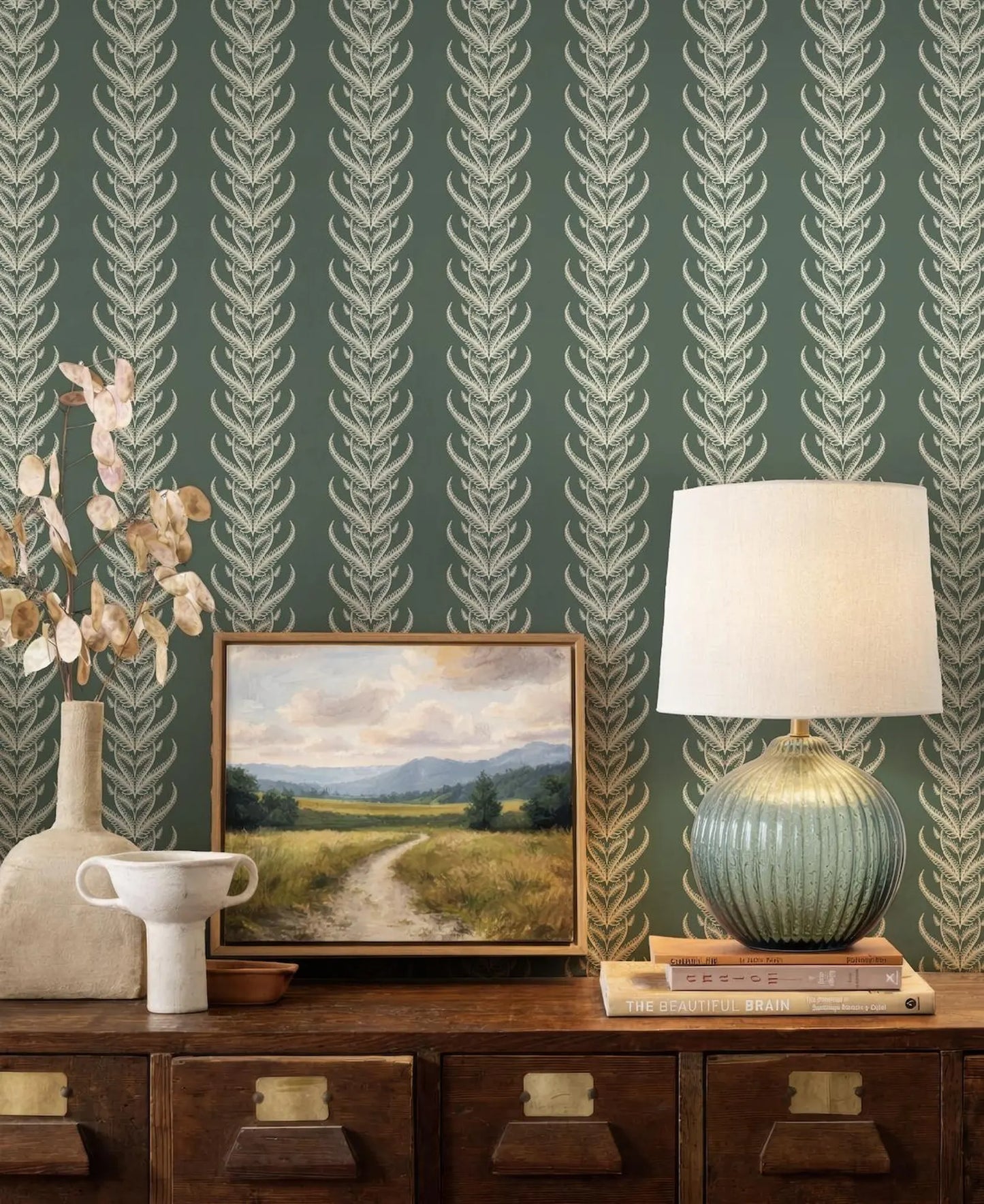 a wooden dresser with a framed landscape painting, a lamp, and a vase on top. The dresser has three drawers and a shelf below it. The wall behind the dresser has a repeating pattern of green and white leaf designs.