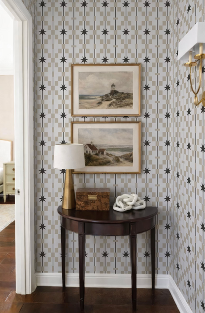 a corner of a room with a wooden table, a lamp, and two framed pictures on the wall above it. The wall has a patterned wallpaper with black stars and a white base.