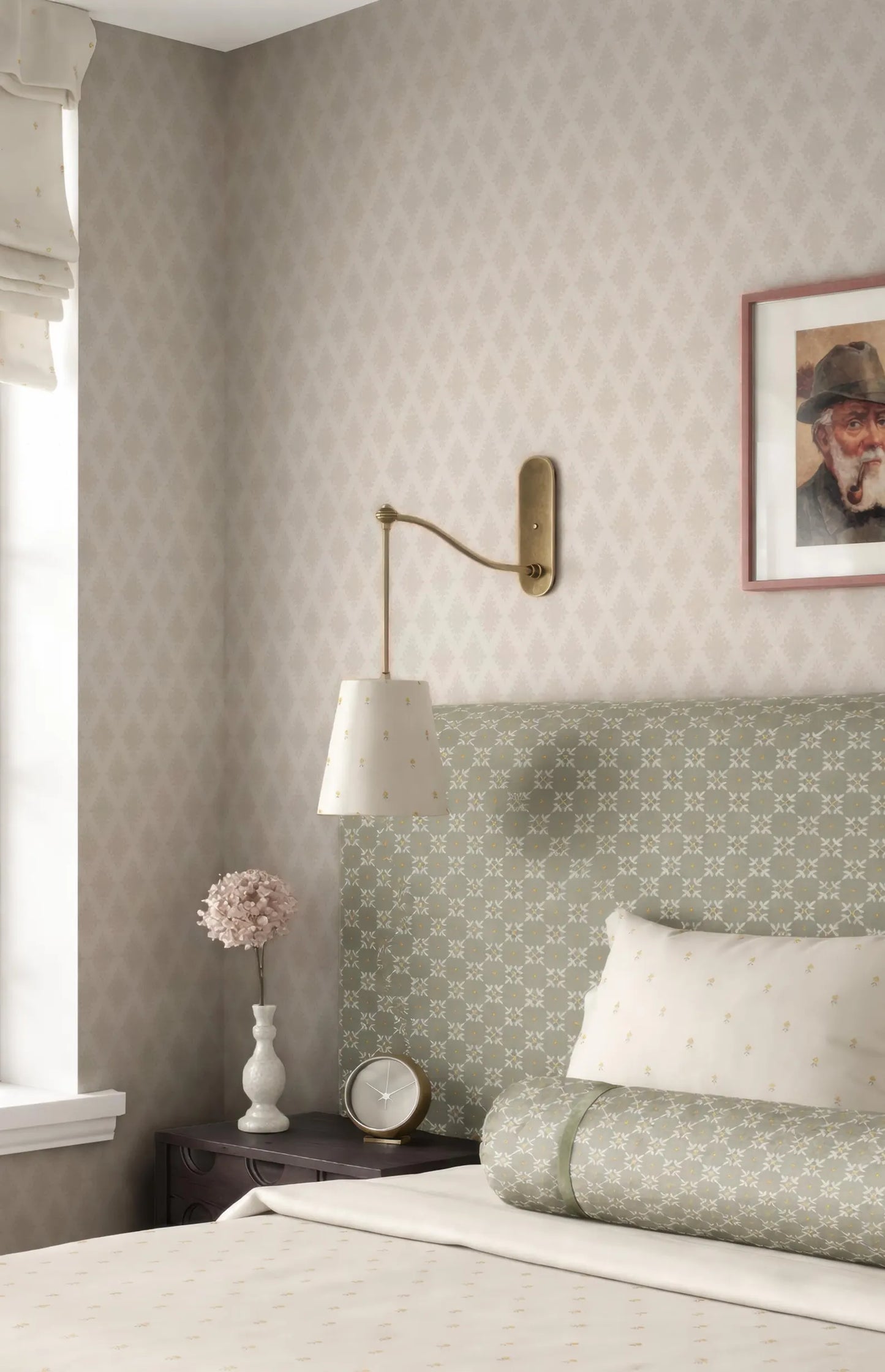 a cozy bedroom with a bed, a nightstand, and a lamp. The bed has a green and white patterned headboard, and there is a framed picture on the wall above it. The nightstand has a white vase with a pink flower, and there is a clock on the nightstand. The lamp is mounted on the wall above the bed.