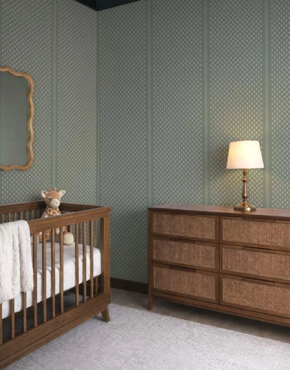 a serene nursery room with a wooden crib, a wooden dresser, and a lamp on the dresser. The room has a soothing green color scheme and a patterned wallpaper.