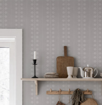 a shelf with various kitchen items, including a wooden cutting board, a white teapot, a white mug, and a stack of plates. The shelf is set against a wall with a gray and white patterned wallpaper.