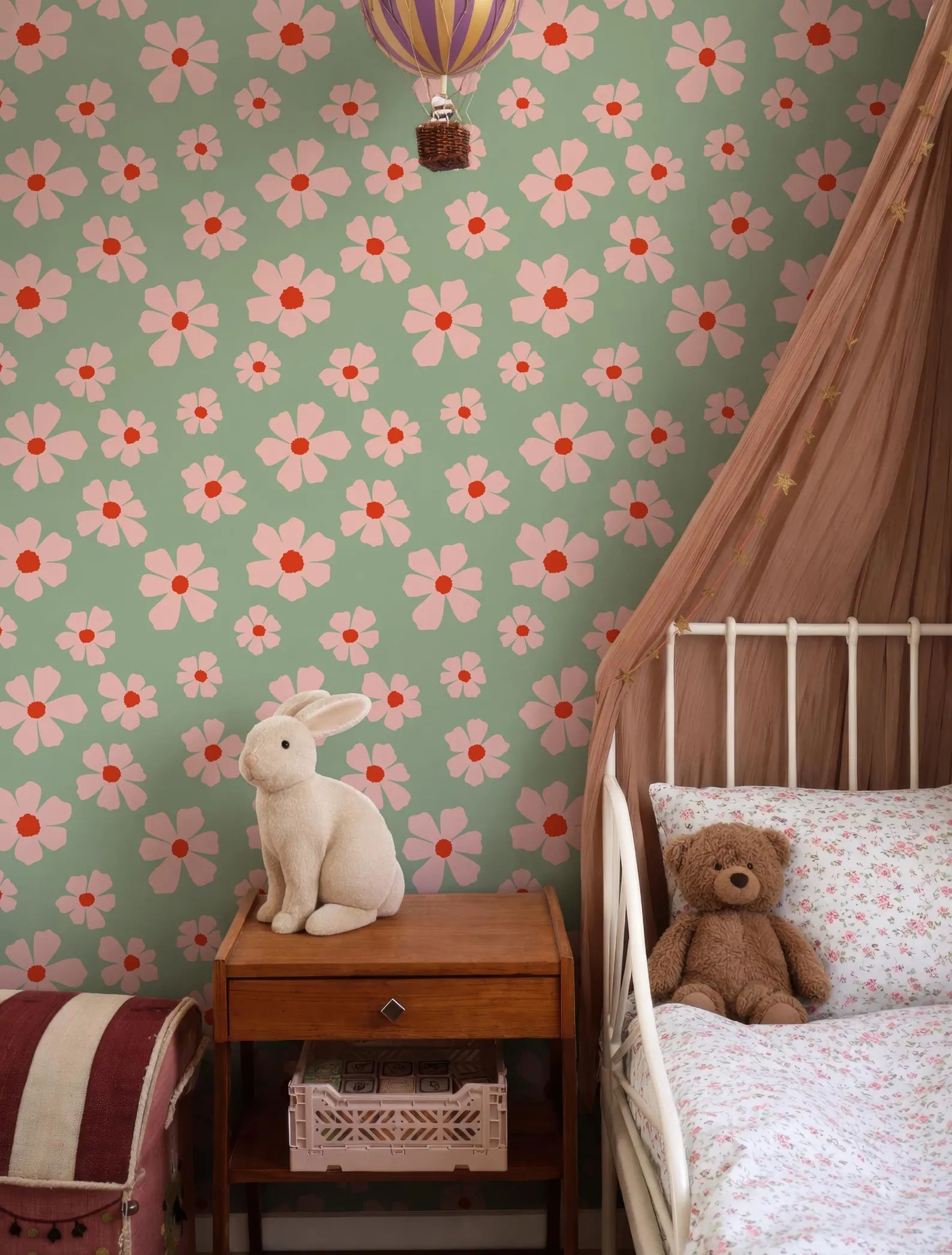 A child's bedroom with a floral wallpaper, a white metal bed frame, a wooden nightstand with a stuffed rabbit, and a pink and white striped backpack.