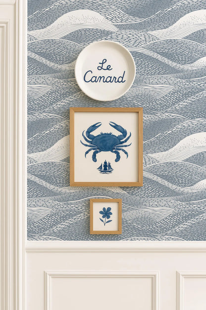 a wall with a blue and white wallpaper pattern, and there are three framed pictures hanging on it. The top picture is a large blue crab, the middle picture is a small blue flower, and the bottom picture is a small blue ship.