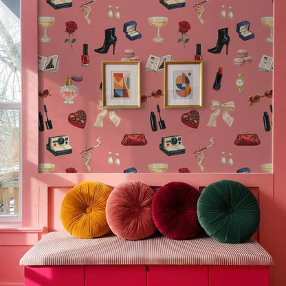 a pink wall with a pattern of various objects, including a rose, a wine glass, a high heel shoe, a camera, and a heart. Above the wall, there are two framed pictures hanging. In front of the wall, there is a pink bench with four colorful pillows on it.