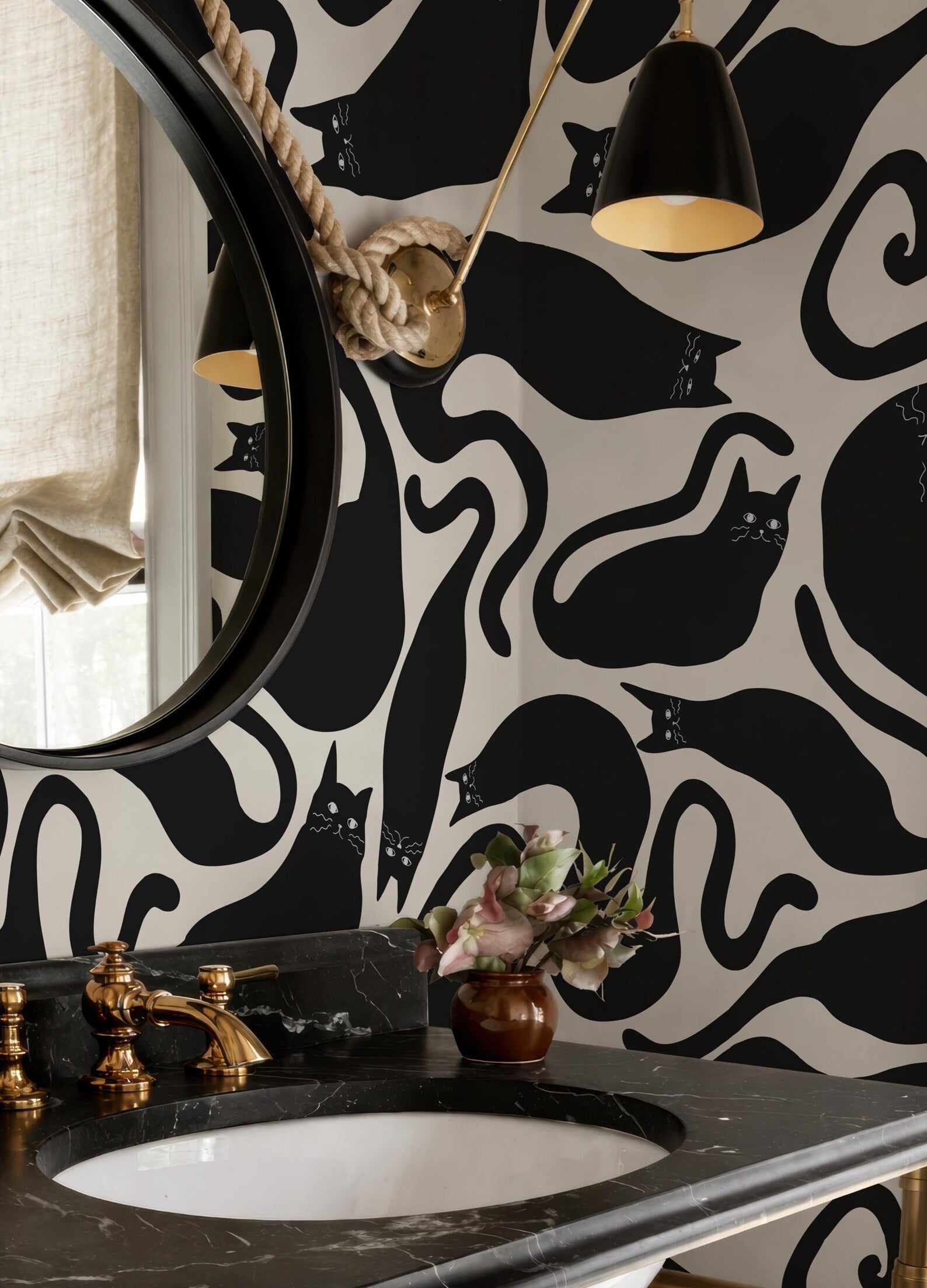 a bathroom with a black and white cat-themed wallpaper, a round mirror, a black vanity with a gold faucet, and a vase of flowers on the counter.