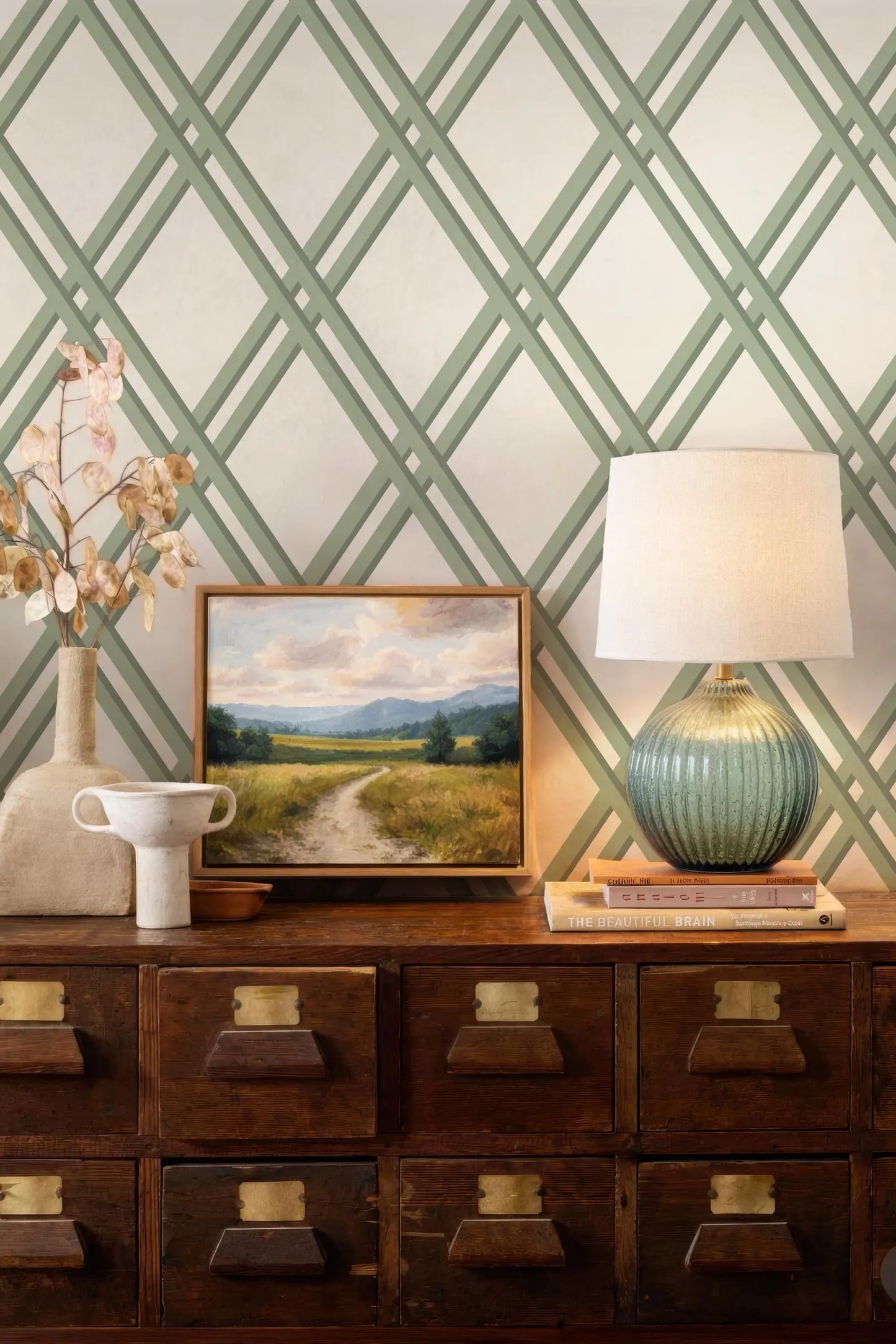a wooden dresser with a painting of a countryside landscape, a lamp, and a vase of flowers on top. The dresser has multiple drawers and a cabinet below it. The wall behind the dresser has a geometric pattern in shades of green and white.