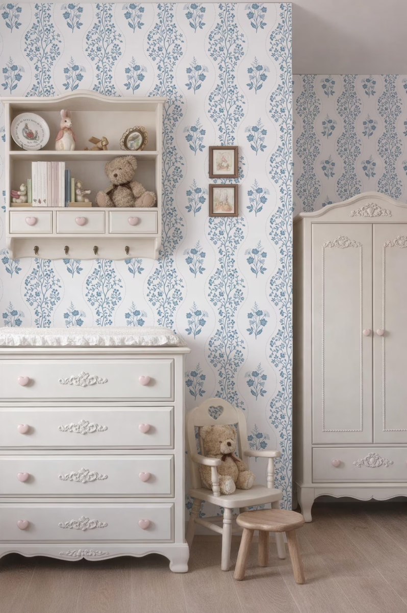 a cozy nursery room with a white dresser, a white chair, and a white cabinet. The walls are covered in a blue floral wallpaper, and there are two framed pictures hanging on the wall. The room also features a teddy bear and a small teddy bear doll, adding a touch of warmth and charm to the space.