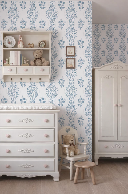 a cozy nursery room with a white dresser, a white chair, and a white cabinet. The walls are covered in a blue floral wallpaper, and there are two framed pictures hanging on the wall. The room also features a teddy bear and a small teddy bear doll, adding a touch of warmth and charm to the space.