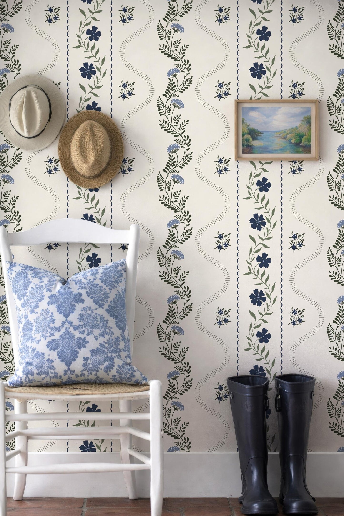 a white chair with a blue and white floral pillow, a pair of black rain boots, and a framed landscape painting on a wall with a floral pattern.