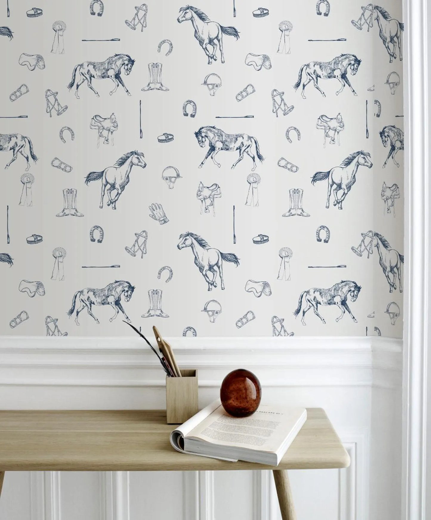 a wallpapered wall with a repeating pattern of horses and various equestrian-related objects, such as boots, helmets, and riding gear. In front of the wall, there is a wooden table with a book, a small wooden box, and a brown ball.