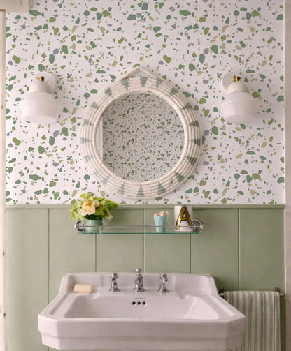 a bathroom with a white sink, a round mirror, and a green and white speckled wall. The bathroom also has a shelf above the sink with a vase of flowers and a letter "A" on it.
