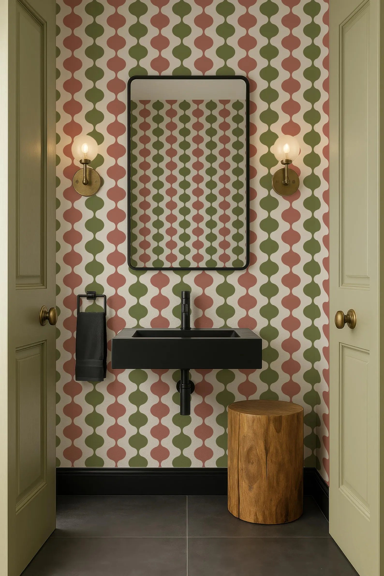 a bathroom with a black sink, a mirror, and two wall sconces. The walls are covered in a patterned wallpaper with a mix of green, red, and white colors. The floor is tiled, and there is a wooden stool in the corner of the room.