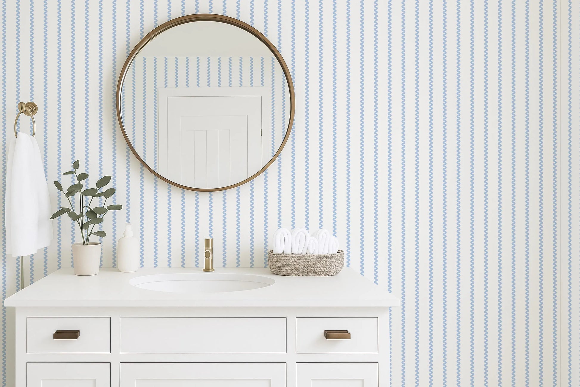 a bathroom with a white sink, a round mirror, and a white cabinet. The bathroom has a striped blue and white wallpaper, and there is a plant on the counter.
