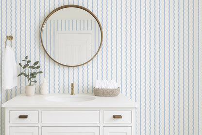a bathroom with a white sink, a round mirror, and a white cabinet. The bathroom has a striped blue and white wallpaper, and there is a plant on the counter.