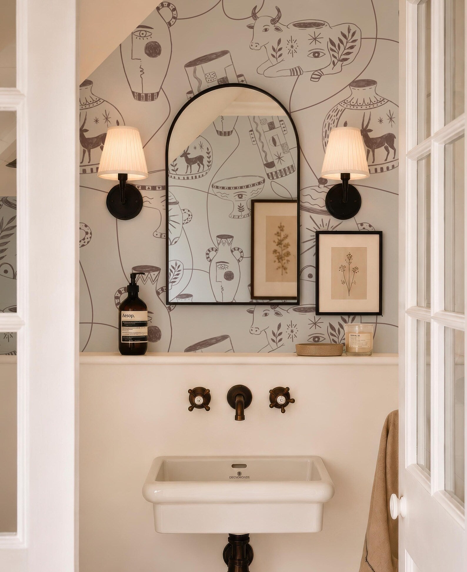 a bathroom with a white sink, a mirror, and two wall sconces. The walls are decorated with a pattern of vases and other decorative elements.