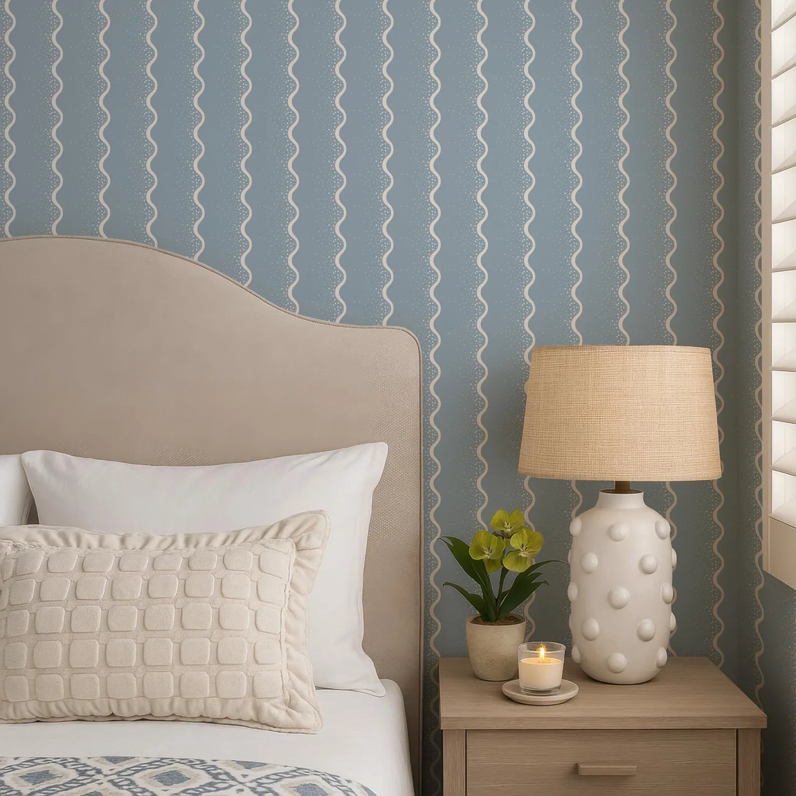 a cozy bedroom scene with a bed, a nightstand, and a lamp. The bed has a beige headboard and white pillows, and the nightstand has a white lamp with a beige shade. There is also a small potted plant and a candle on the nightstand.