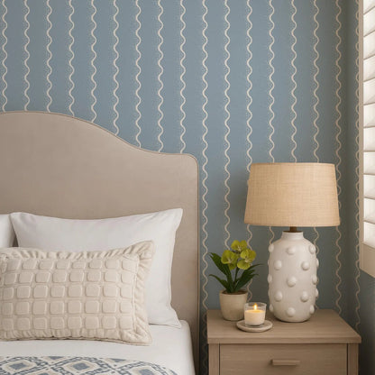 a cozy bedroom scene with a bed, a nightstand, and a lamp. The bed has a beige headboard and white pillows, and the nightstand has a white lamp with a beige shade. There is also a small potted plant and a candle on the nightstand.