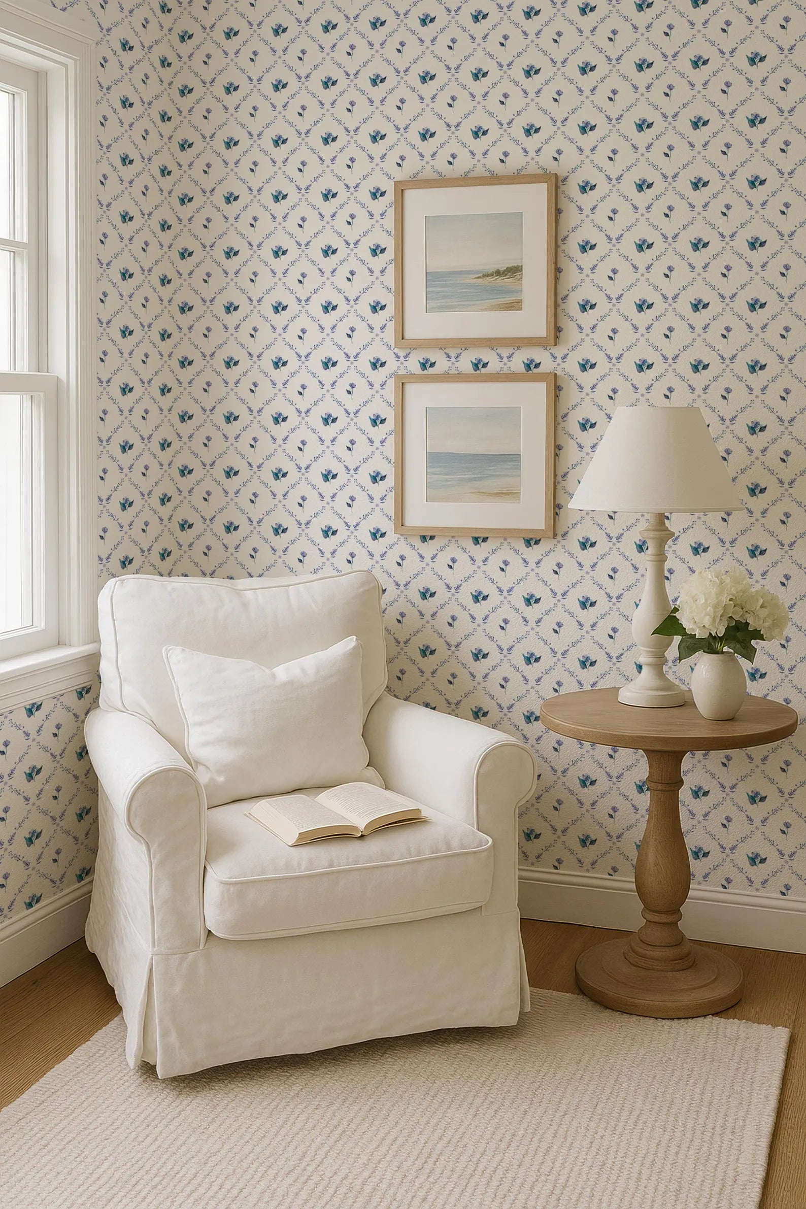 a cozy corner of a room with a white armchair, a small side table with a lamp and a vase of flowers, and two framed pictures on the wall.