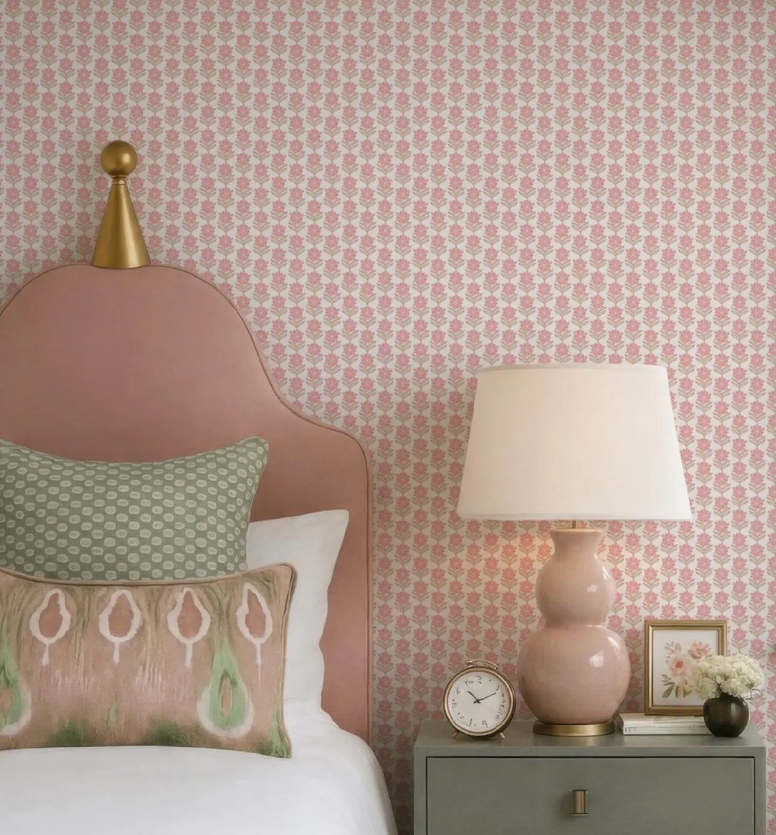 a bedroom with a pink headboard, a white bedspread, and a pink lamp on a nightstand. The wall behind the bed has a pink floral wallpaper.