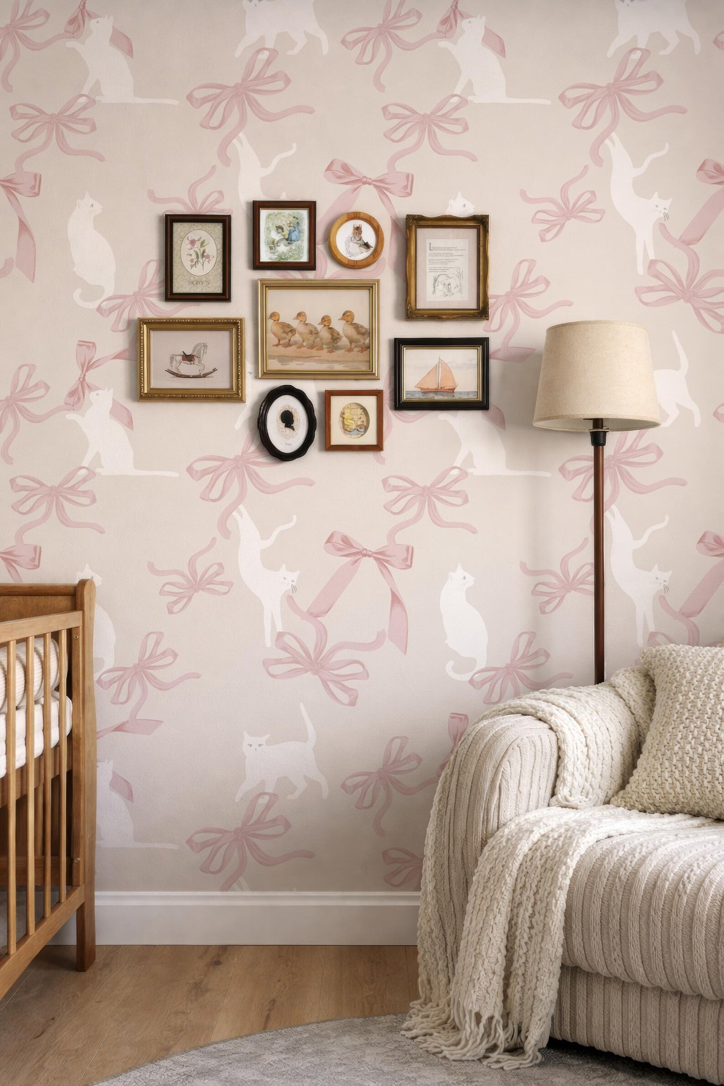 a cozy and charming nursery room with a pink and white color scheme. The walls are adorned with a pattern of pink bows, and there is a wooden crib on the left side of the room. A white sofa is positioned on the right side, and a lamp is placed on the floor next to the sofa. The room also contains a collection of framed pictures and a cat illustration, adding a personal touch to the space.