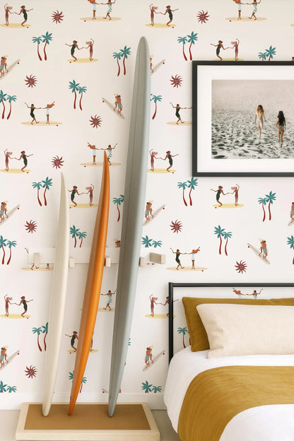 a bedroom with a wallpapered wall that has a repeating pattern of surfers and palm trees. There is a bed with a yellow blanket and a framed photo of a beach scene above it. Two surfboards are leaning against the wall, and a large orange surfboard is propped up against the wall.