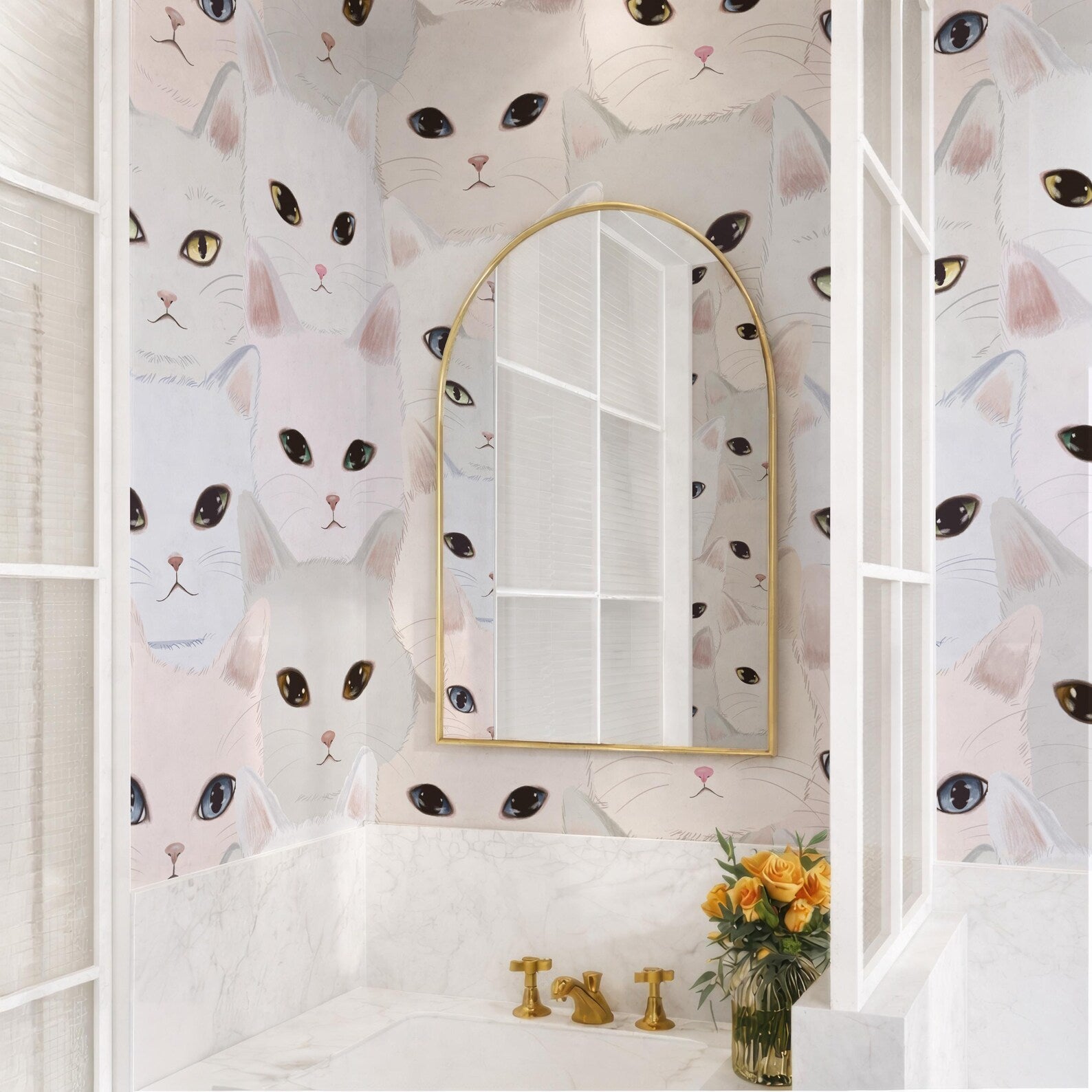 a bathroom with a white sink and a gold faucet, and a mirror above it. The wall behind the sink is covered in a repeating pattern of cat faces in various colors and sizes. There is also a vase of flowers on the counter next to the sink.