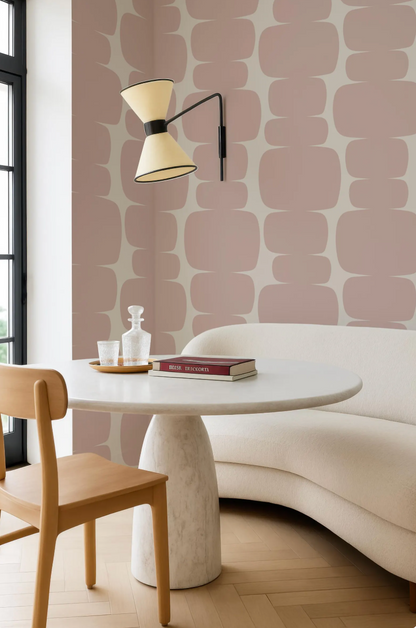 a modern and minimalist living space with a white sofa, a round white table, and a wooden chair. The walls are adorned with a geometric pattern in shades of pink and white, creating a visually appealing and cohesive design. A lamp is mounted on the wall, providing soft lighting to the room.