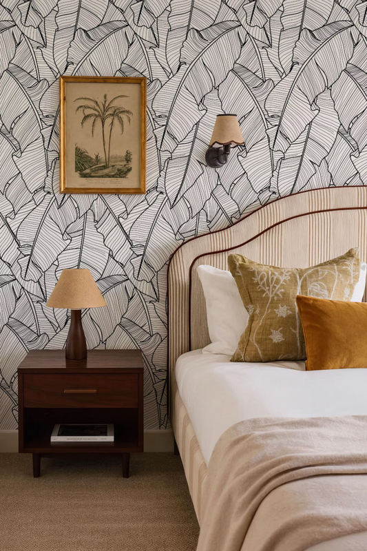 a bedroom with a bed, nightstand, and wall decor. The bed has a striped headboard and is adorned with decorative pillows. The nightstand has a lamp on it, and there is a framed picture hanging on the wall above the bed. The wall is covered in a patterned wallpaper with a tropical leaf design.