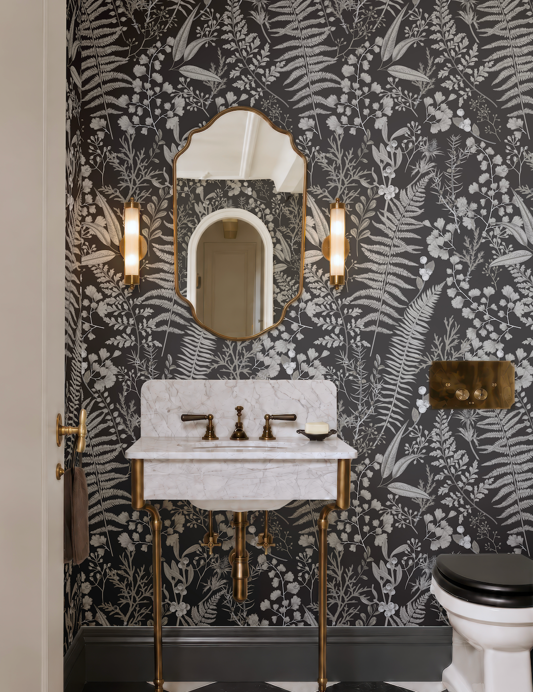 a bathroom with a white marble sink, a gold faucet, and a black and white floral wallpaper. The bathroom also includes a toilet and two wall sconces.