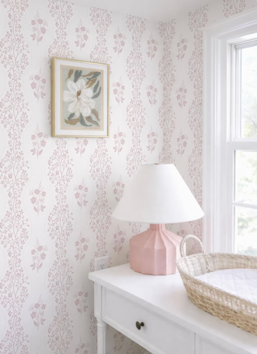 a white dresser with a pink lamp and a woven basket on top, set against a wall with a floral wallpaper.