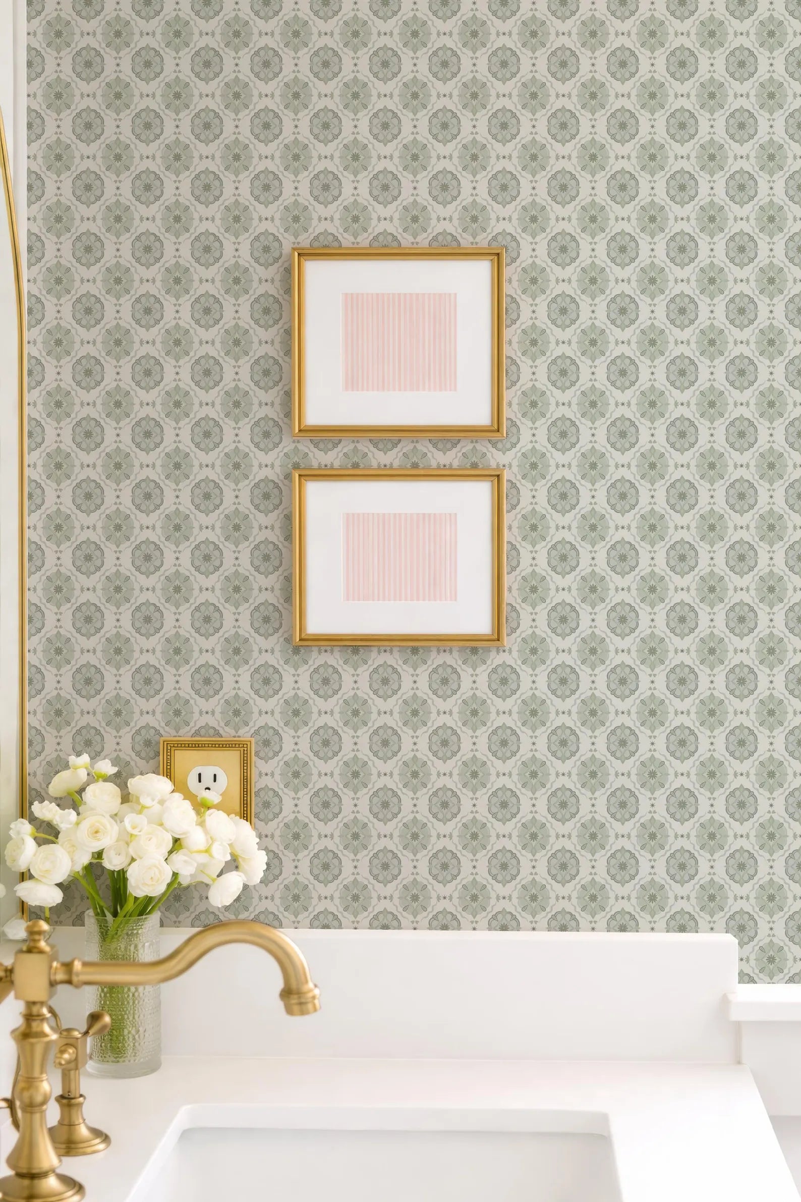 a bathroom with a white sink, a gold faucet, and a vase of white flowers. Above the sink, there are two framed pictures hanging on the wall.
