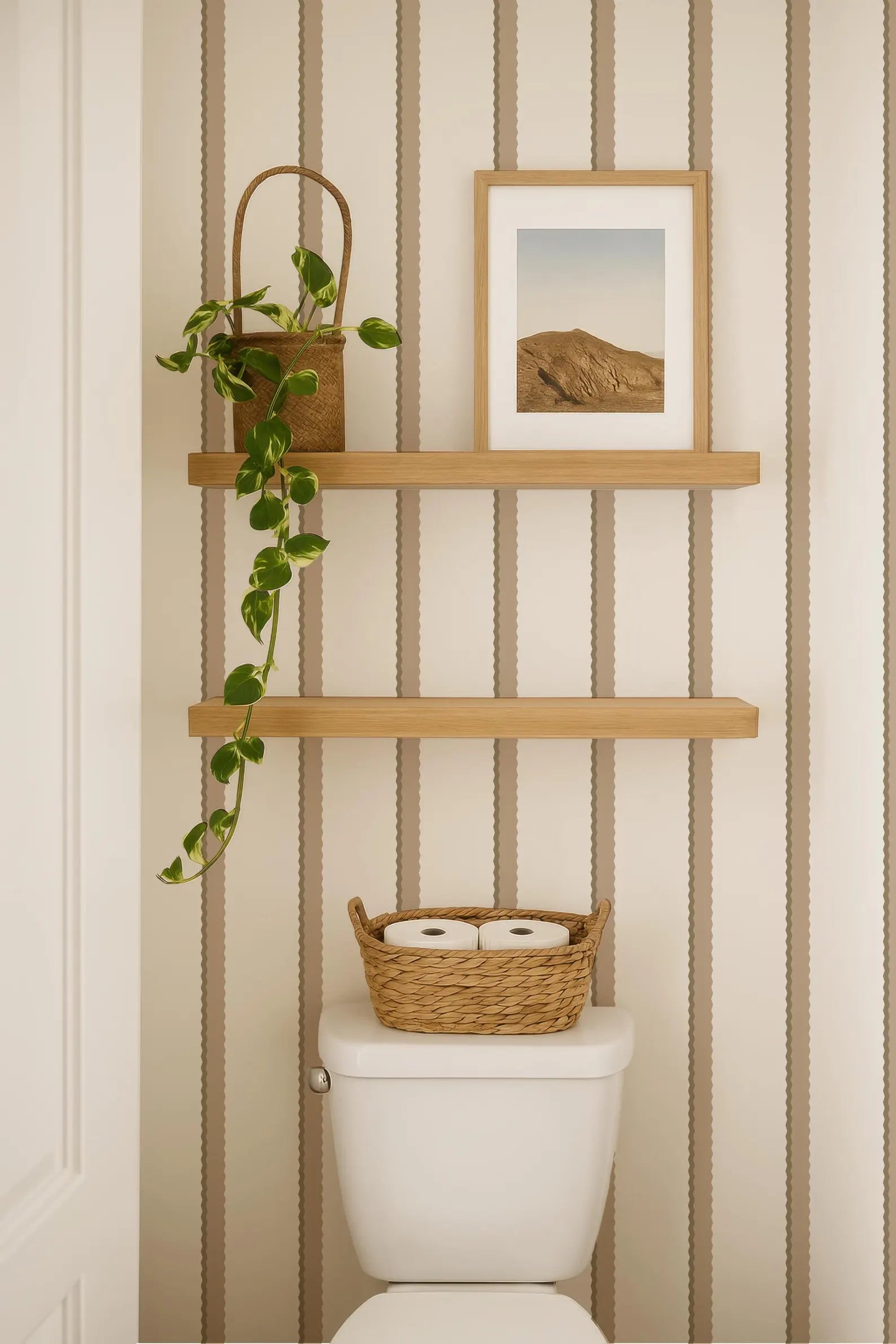 a bathroom with a toilet, a wooden shelf above it, and a framed picture on the shelf. The bathroom has a striped wallpaper, and there is a basket of toilet paper on the tank of the toilet.
