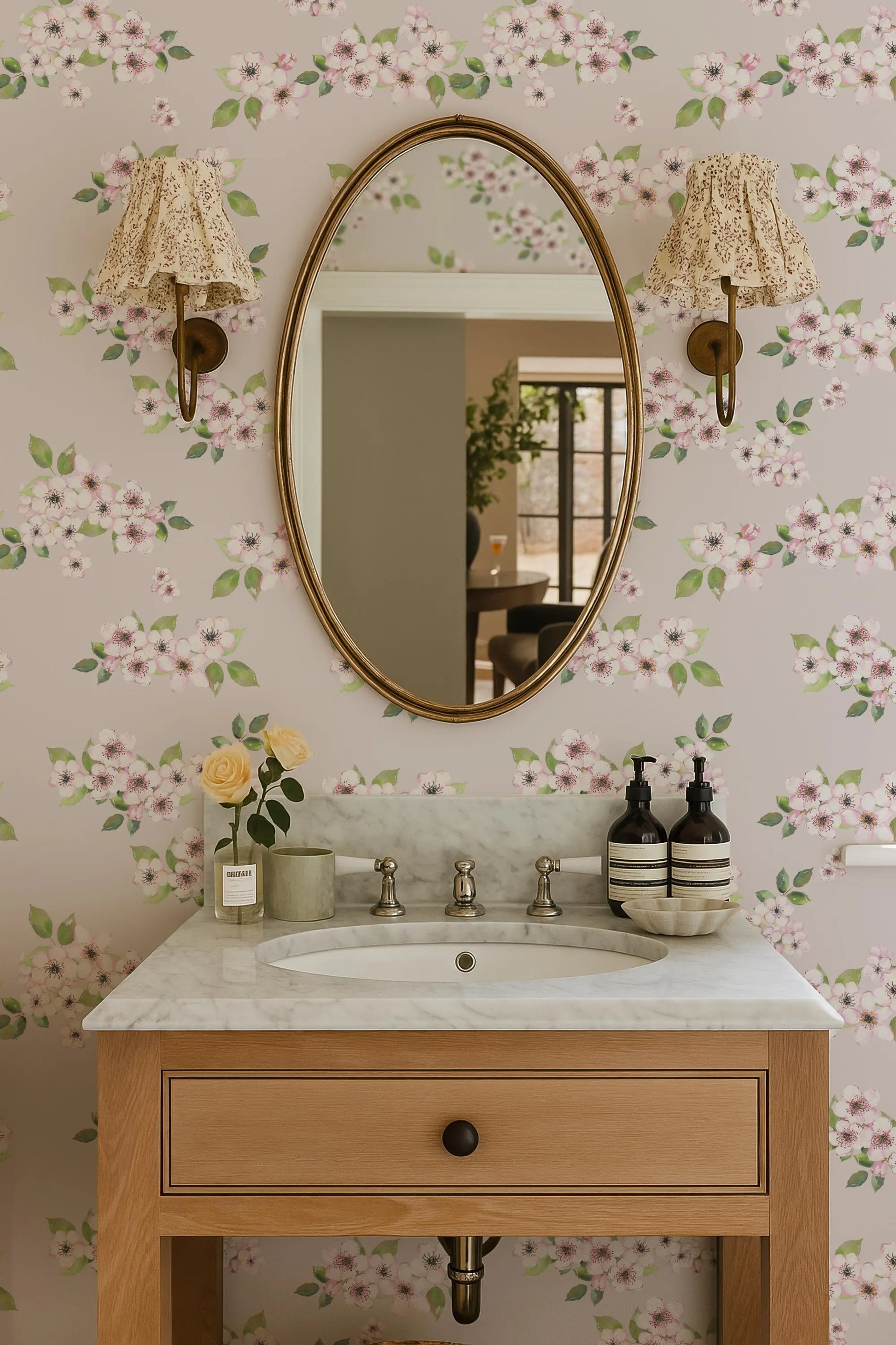 a bathroom sink with a marble countertop, a large oval mirror, and two wall sconces. The walls are decorated with a floral wallpaper pattern, and there are some decorative elements such as a vase of flowers and a bottle on the countertop.