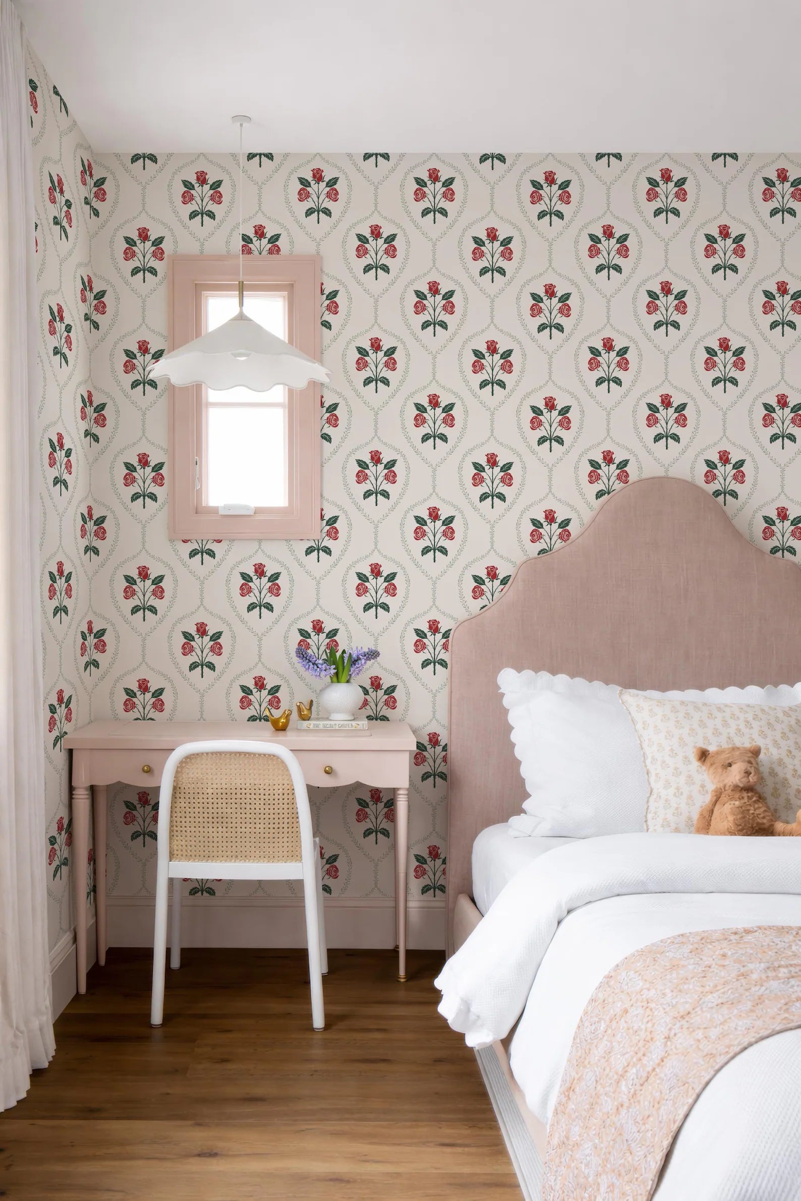 a cozy bedroom with a pink headboard, floral wallpaper, and a white desk with a vase of flowers. The room has a warm and inviting atmosphere, with a comfortable bed and a chair.