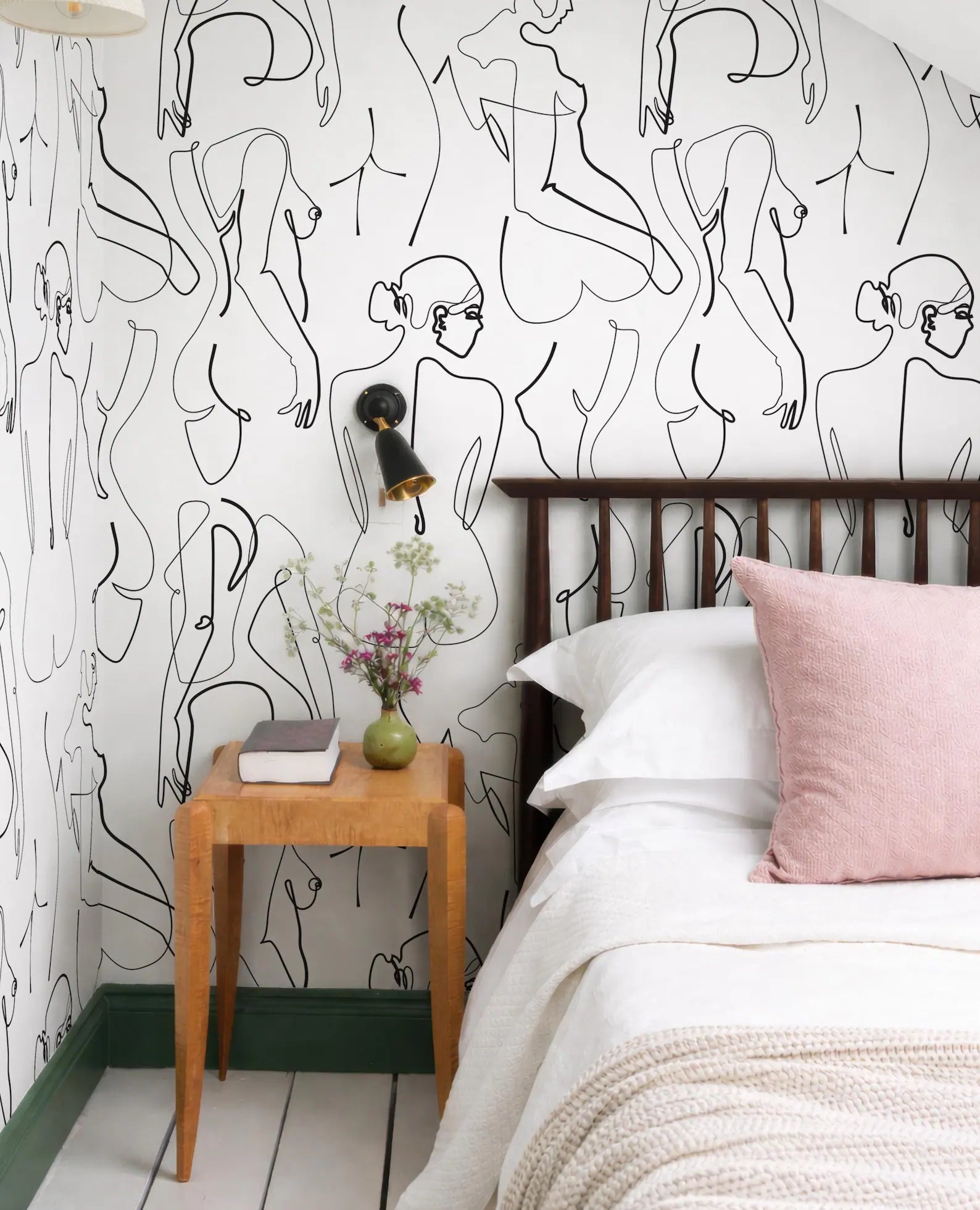 a bedroom with a bed, a wooden nightstand, and a wallpapered wall with abstract line drawings of human figures.