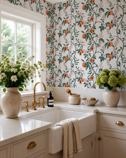 a well-decorated kitchen with a white sink, a vase of flowers, and a bowl of fruit. The wall behind the sink is covered in a floral wallpaper with orange and green accents.