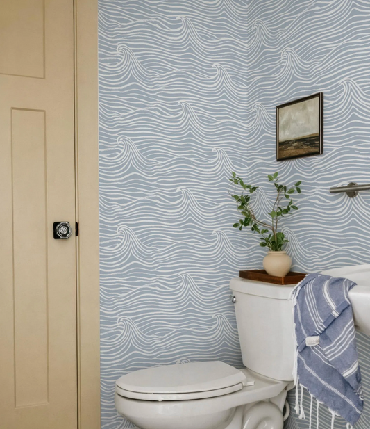 a bathroom with a white toilet, a towel hanging on the side, and a framed picture on the wall. The walls are covered in a blue and white wave-patterned wallpaper, and there is a plant on the toilet tank.
