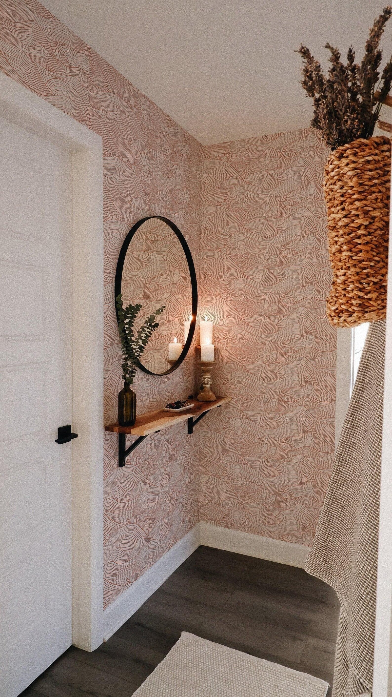 a cozy corner of a room with a mirror, a shelf with candles, and a woven basket with dried flowers. The walls are covered in a pink and white patterned wallpaper, and the floor is made of wood.