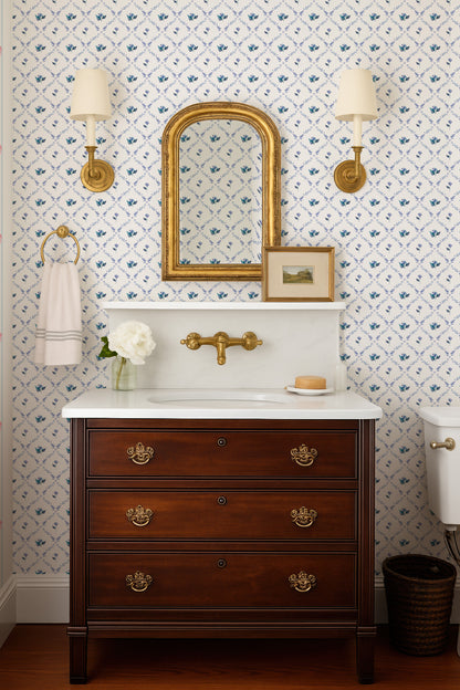 a bathroom with a wooden vanity, a white sink, and a mirror above it. The walls are decorated with blue and white floral wallpaper, and there are two wall sconces on either side of the mirror. The vanity has three drawers and a white countertop.