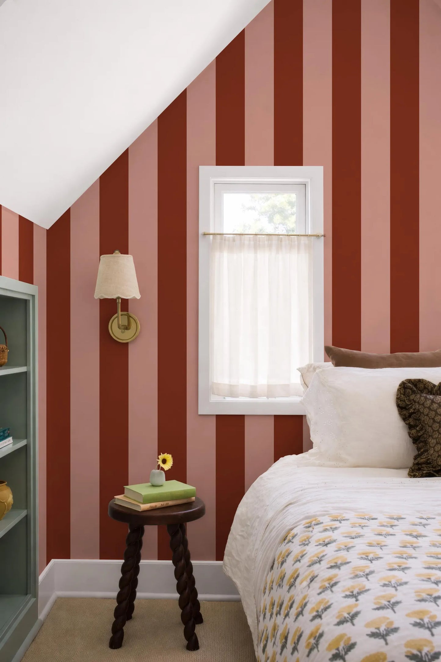 a cozy bedroom with a bed, a small table, and a window with a white curtain. The walls are painted in a striped pattern in shades of red and pink, and the room has a warm and inviting atmosphere.