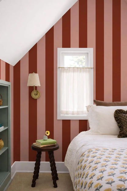 a cozy bedroom with a bed, a small table, and a window with a white curtain. The walls are painted in a striped pattern in shades of red and pink, and the room has a warm and inviting atmosphere.