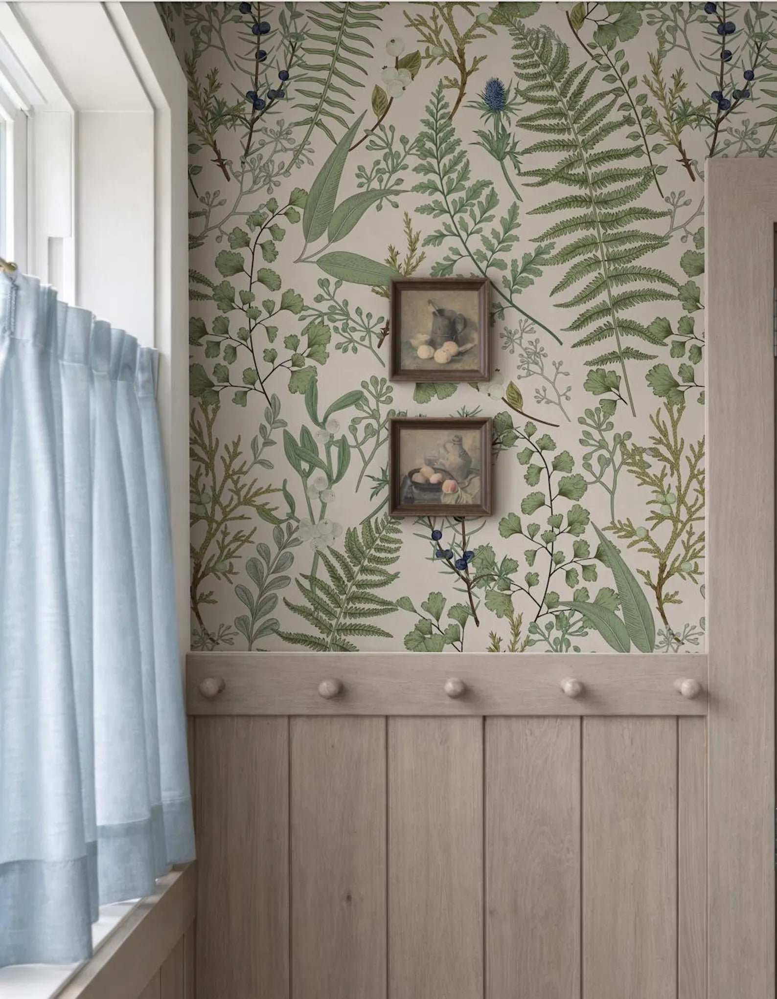 a wall with a green and white floral wallpaper, and two framed pictures hanging on it. The wall is also adorned with a wooden panel and a window with light blue curtains.