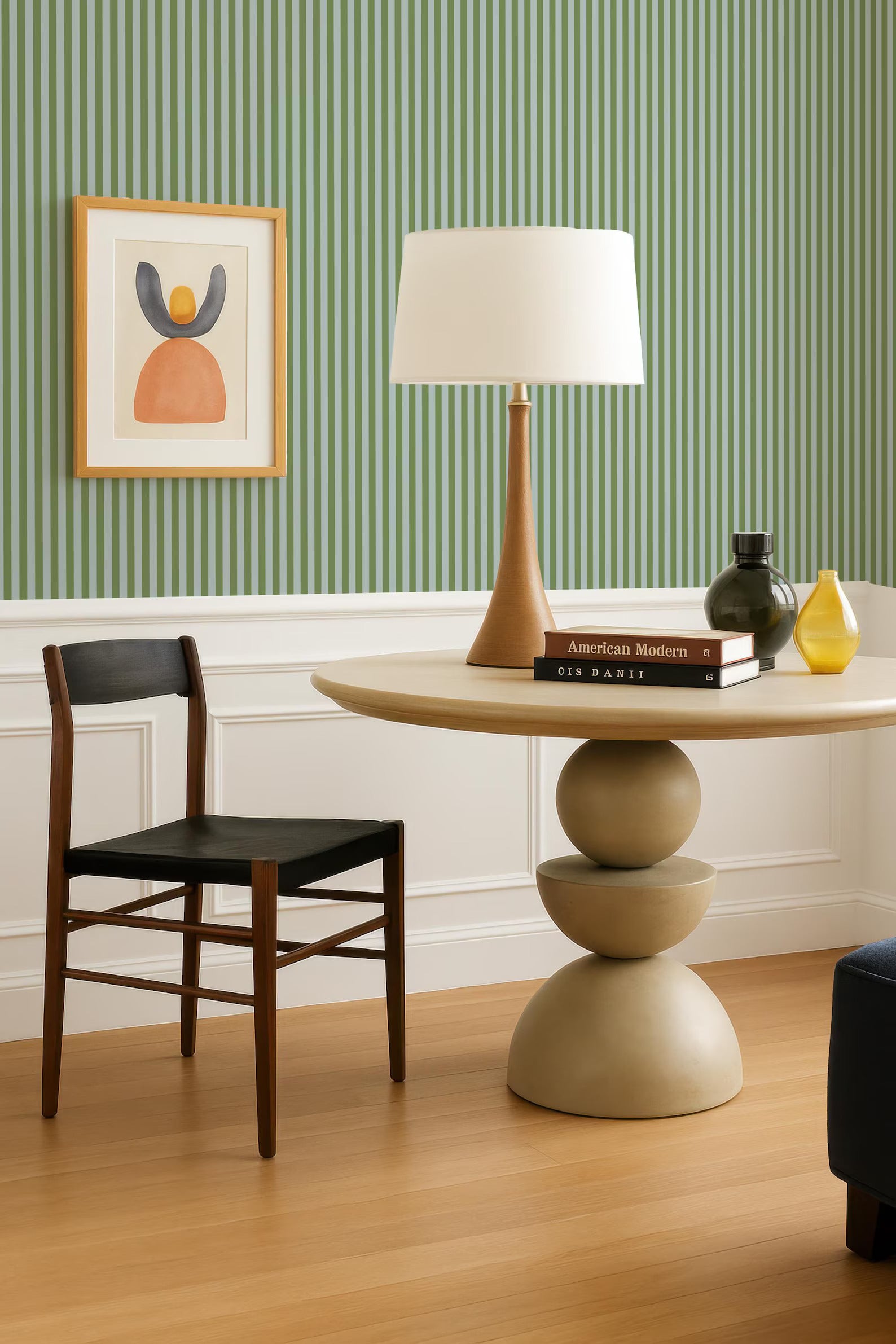 a modern living room with a wooden chair, a round table with a lamp, and a framed artwork on the wall.
