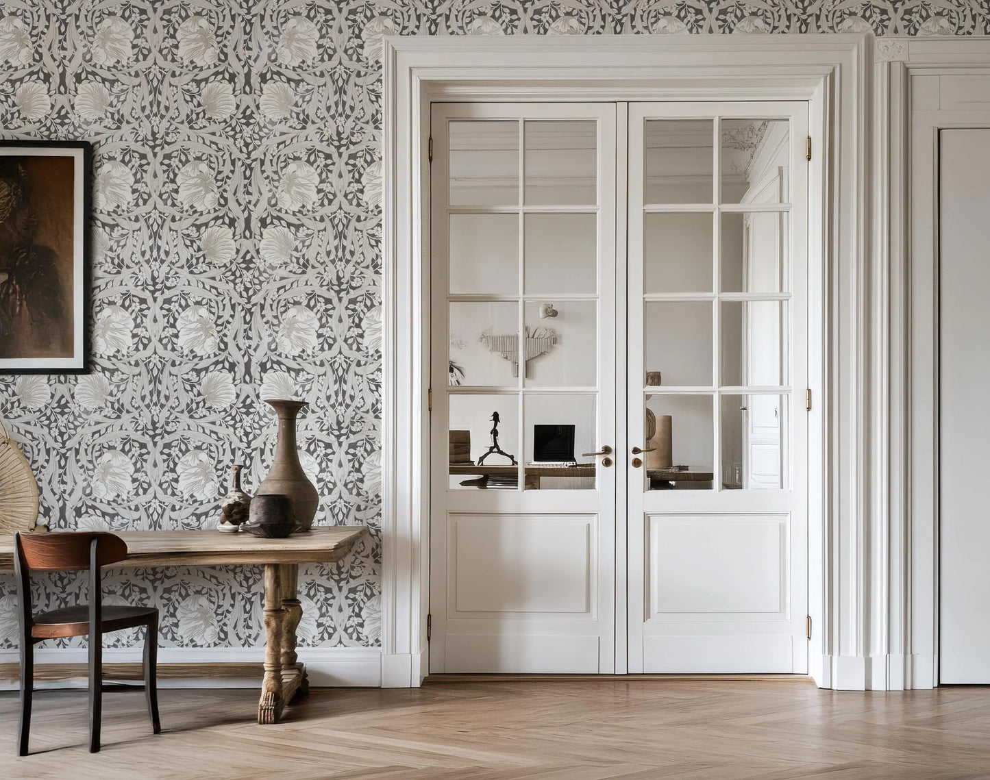 a room with a wooden table, a vase, and a chair. The room has a patterned wallpaper and a large window with white curtains.