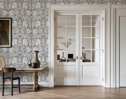 a room with a wooden table, a vase, and a chair. The room has a patterned wallpaper and a large window with white curtains.