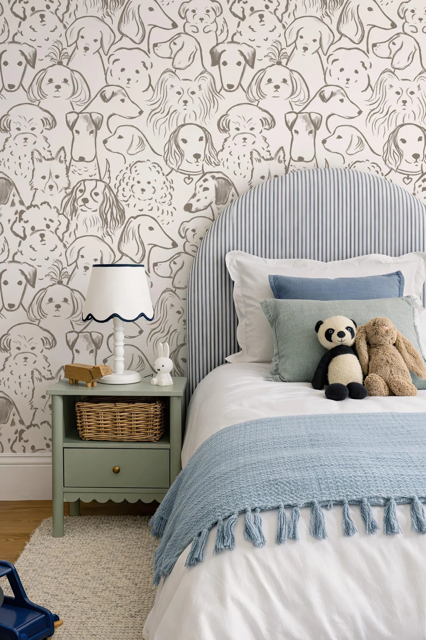 a cozy bedroom scene with a bed, nightstand, and stuffed animals. The bed has a striped headboard and is adorned with a blue blanket and a panda stuffed animal. The nightstand has a white lamp and a small basket. The wall behind the bed is decorated with a repeating pattern of dog faces in various shades of gray and brown.