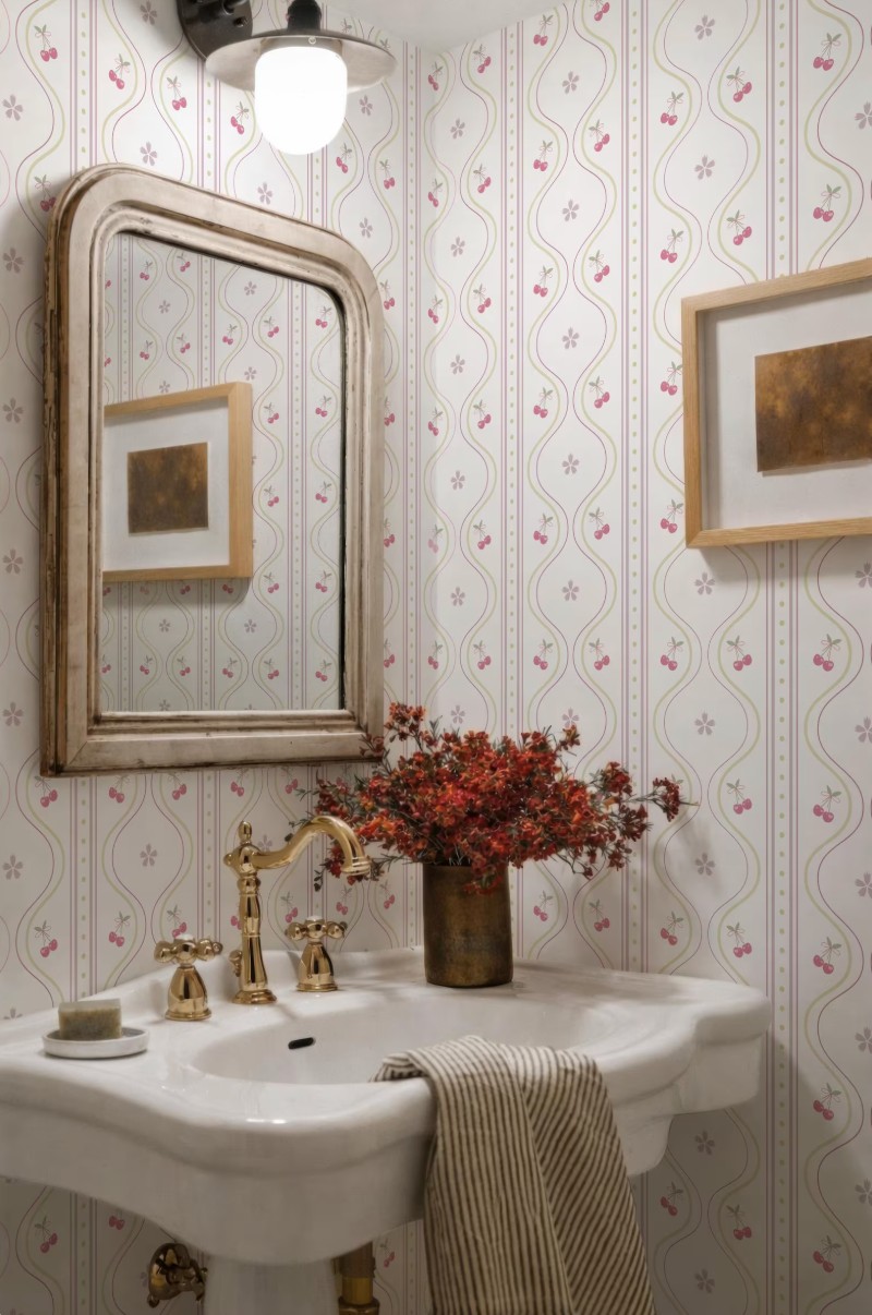 a bathroom with a white sink, a gold faucet, and a framed picture on the wall. The walls are covered in a patterned wallpaper with a repeating design of pink and white flowers. A vase of red flowers is placed on the sink, adding a touch of color and natural beauty to the space.