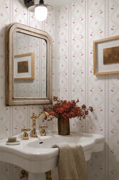 a bathroom with a white sink, a gold faucet, and a framed picture on the wall. The walls are covered in a patterned wallpaper with a repeating design of pink and white flowers. A vase of red flowers is placed on the sink, adding a touch of color and natural beauty to the space.