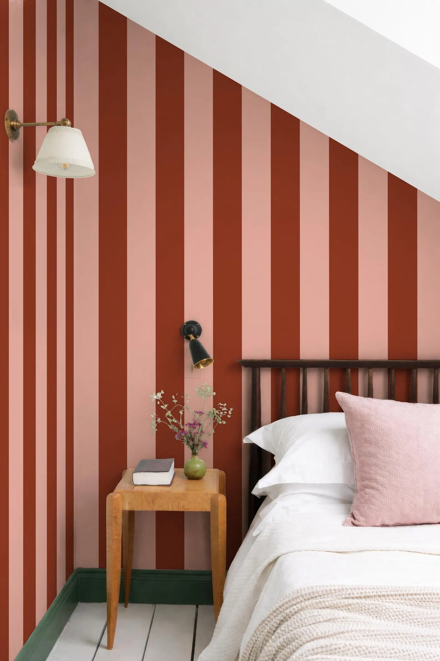 a bedroom with a bed, a wooden nightstand, and a striped wall behind the bed. The wall has vertical stripes in shades of red and pink, creating a vibrant and cozy atmosphere. The room also includes a lamp, a vase with flowers, and a book on the nightstand, adding to the overall aesthetic and comfort of the space.