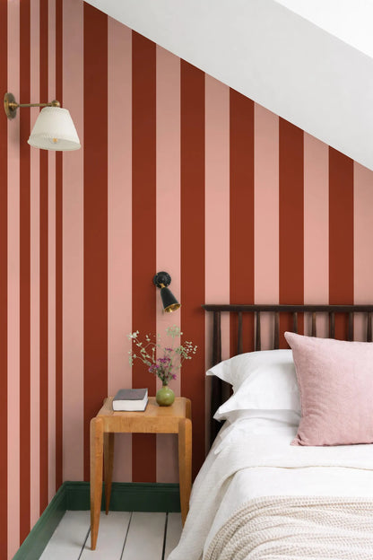 a bedroom with a bed, a wooden nightstand, and a striped wall behind the bed. The wall has vertical stripes in shades of red and pink, creating a vibrant and cozy atmosphere. The room also includes a lamp, a vase with flowers, and a book on the nightstand, adding to the overall aesthetic and comfort of the space.